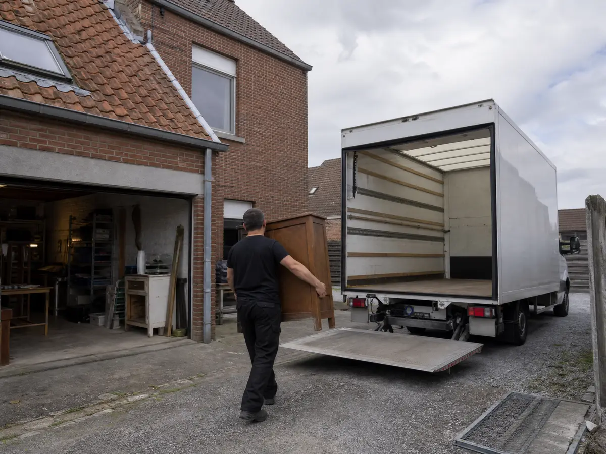 Crew member carries a cabinet from a garage workshop to a box truck during a house clearance in Roeselare
